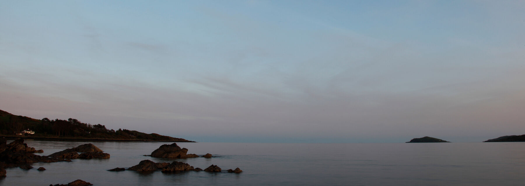 The sea at Rockcliffe seen at dusk, with the moon in the sky. Rocks run into the still, almost purple-blue water.
