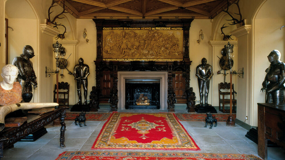 A large entrance hall in a castle with a flagstone floor. Large red and orange rugs cover the middle. Suits of armour stand around the side of the room, and in the centre is a large fireplace with a large plaster decoration above.