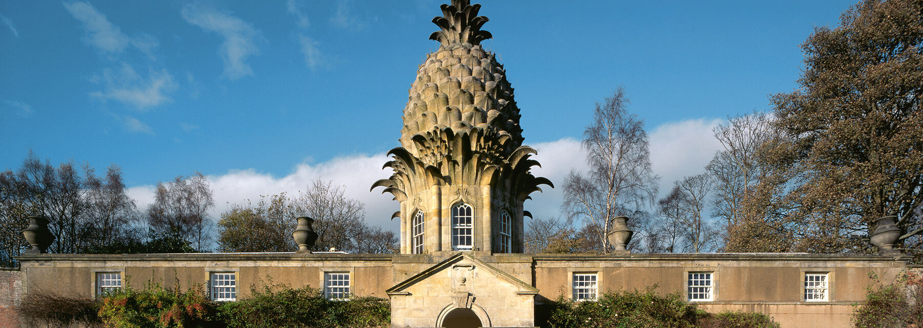 A pineapple-shaped building stands at the centre of a tall, stone garden wall. Shrubs are trained against the garden wall. Tall trees stand at either edge.