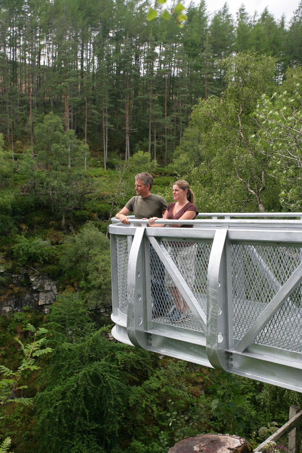 A man and a woman stand on a metal cantilevered viewing platform that sticks out over a gorge. Green trees cover the steep gorge sides.