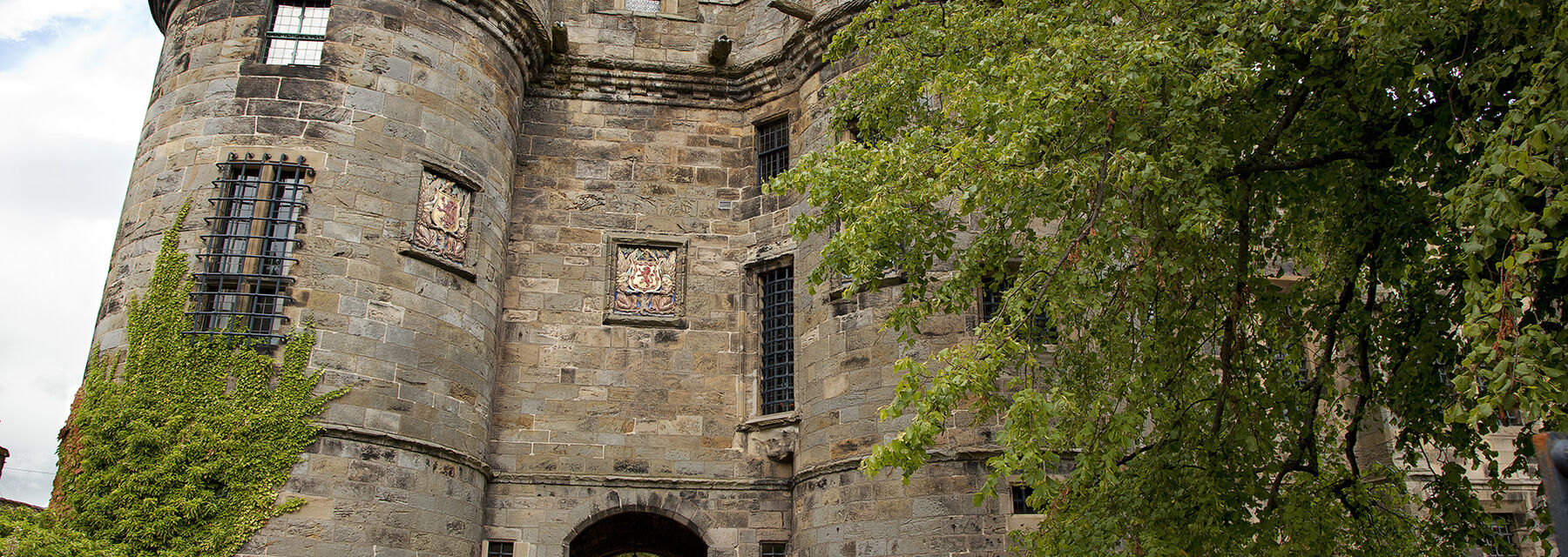 A close-up view of the entrance arch to Falkland Palace, with a round tower to the left. Large green leafy trees stand either side of the entrance path.