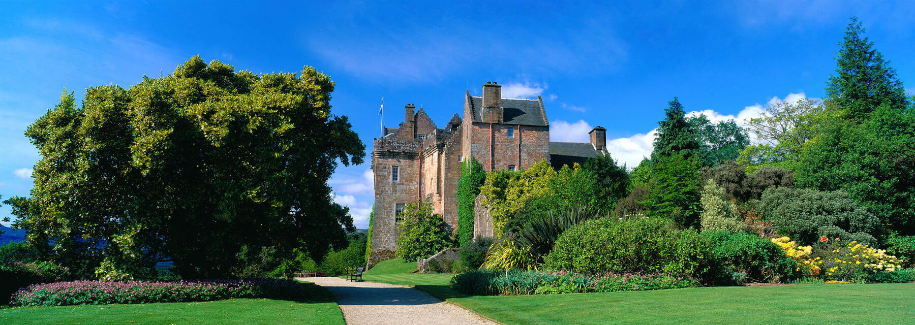 A view of Brodick Castle from the entrance gate, on a bright sunny day with blue sky above. Tall green trees stand either side of the path that leads to the castle. On the right is a bed of established shrubs, many in flower.