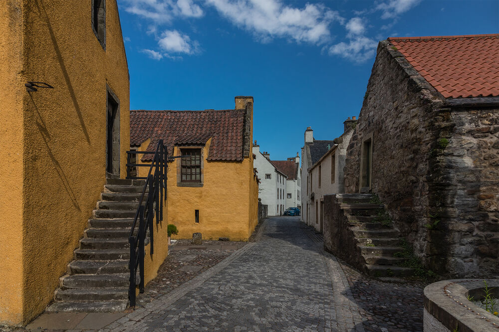 A narrow cobbled street passes between old buildings in Culross. On the left is a 17th-century house with bright yellow walls and red tiles on the roof. An external stone staircase leads up to the first floor.