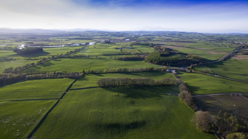 An aerial view of farm fields, divided by hedgerows, pockets of woodland and a couple of roads. Occasional white buildings can be seen in the landscape. A river winds in the distance.