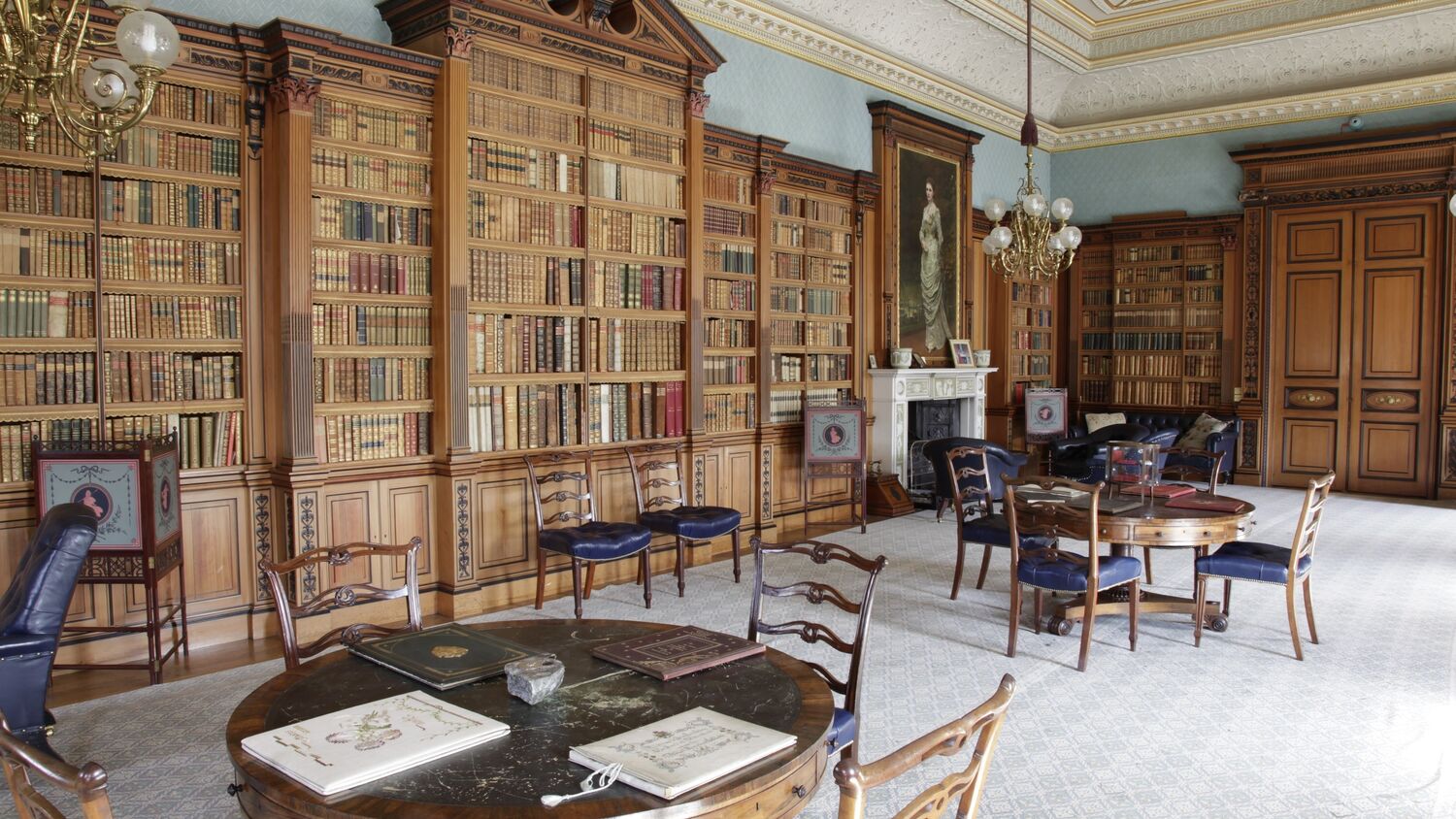 The library at Haddo House, a large and light room with bookshelves from the floor to the ceiling along the walls, low hanging chandeliers, and tables and chairs. There is a portrait of a woman in a long dress hung above a fireplace.