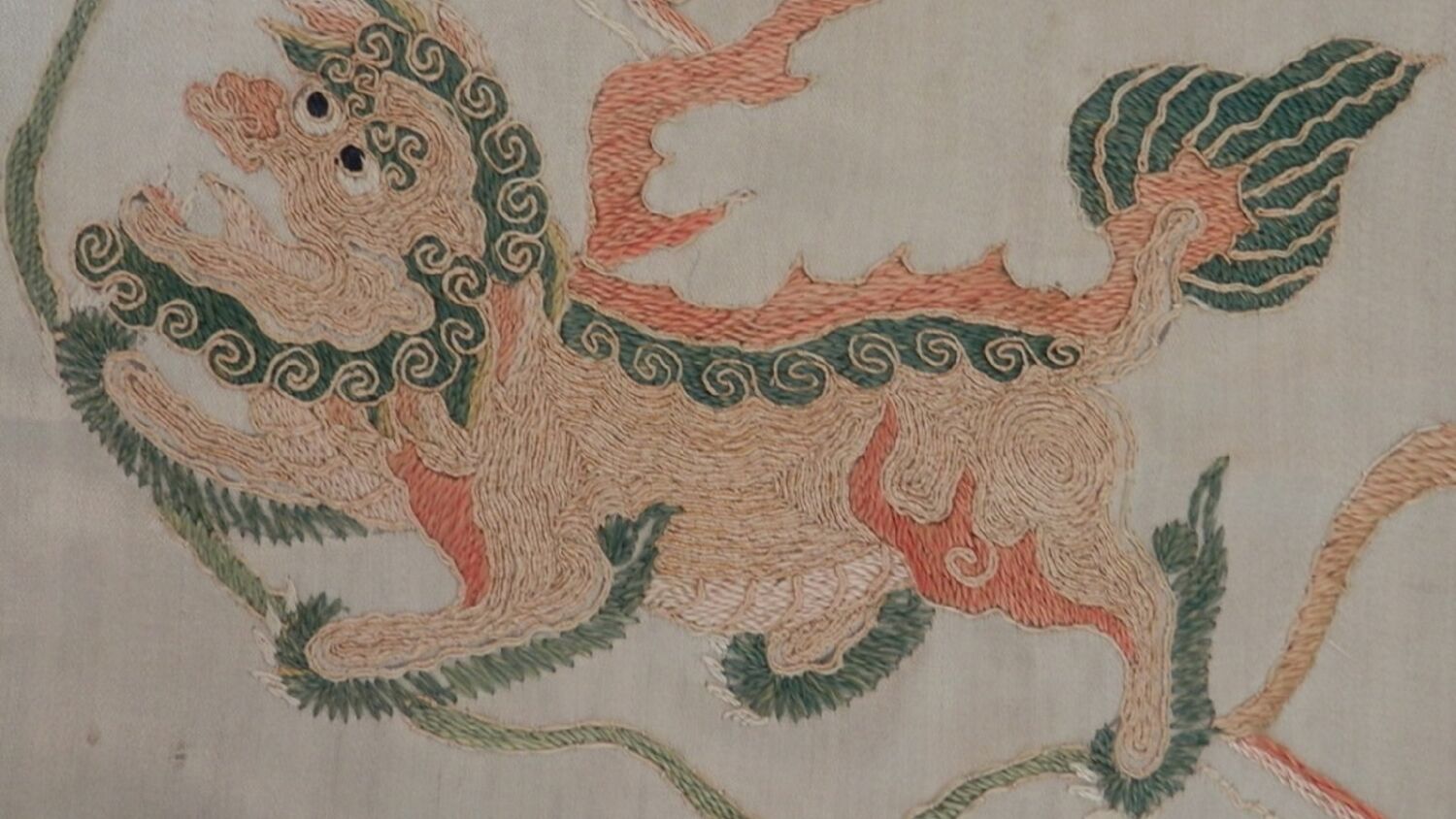 A close-up image of an embroidered Chinese-style lion on a silk panel. The lion is sewn in green, red and orange thread.