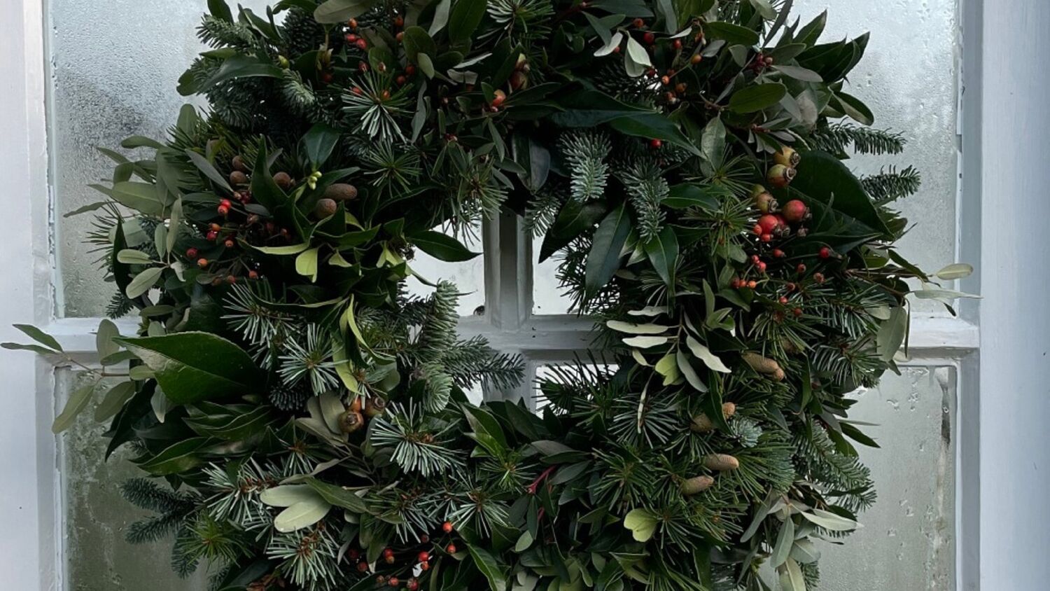 A close-up view of a wreath hanging on a white door with glass panes. The wreath is made mostly from evergreen foliage, as well as some cones and berries.