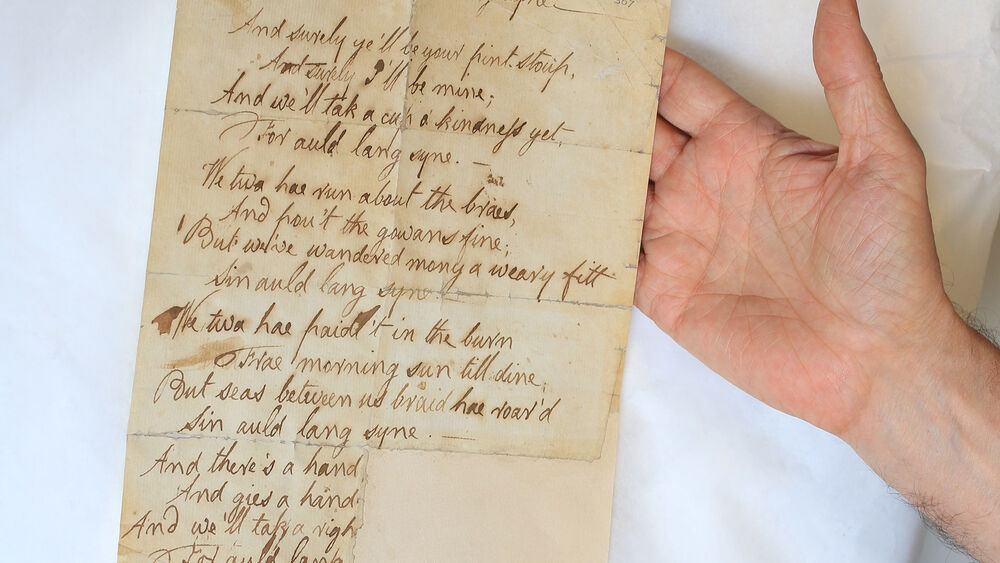 Two hands hold out a page of Burns's handwritten manuscript for 'Auld lang syne'.