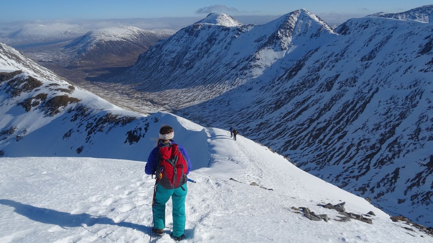 A woman dressed in full winter gear stands almost at the summit of a snow-covered mountain, looking down towards a ridge path, with the glen far below. The sky is bright blue and the snow is glistening.