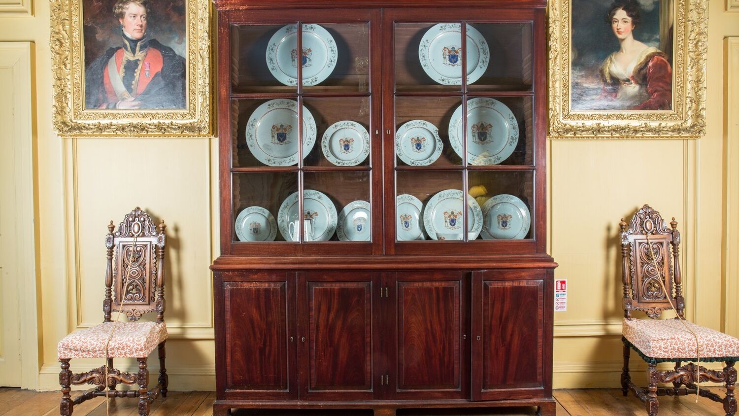 A large wooden bookcase with glass doors at the top. The shelves are filled with china plates instead of books. A chair with a decorative carved back stands either side of the bookcase.