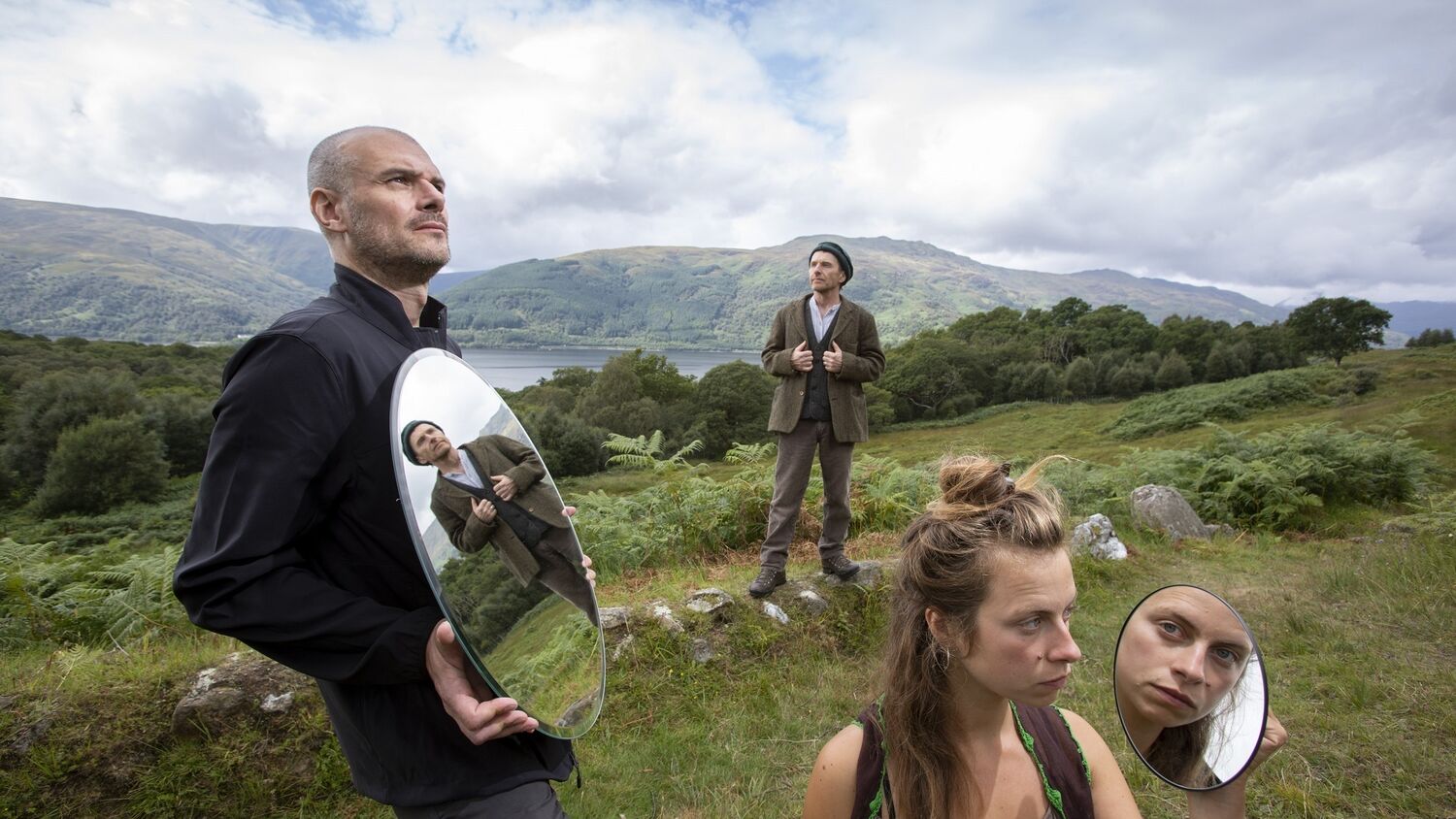 Three people stand on a grassy hillside overlooking Loch Lomond. The man on the left holds an oval mirror, which shows the reflection of the man in the middle, holding the lapels of his tweed jacket. The lady on the right holds a circular mirror to show a reflection of her face.