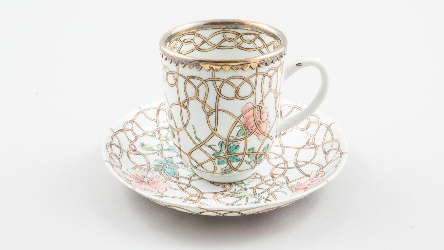 A tea cup sitting on a matching saucer is displayed against a plain grey background. It has a silver-gilt mount and is decorated with pink flowers and gold tendrils.