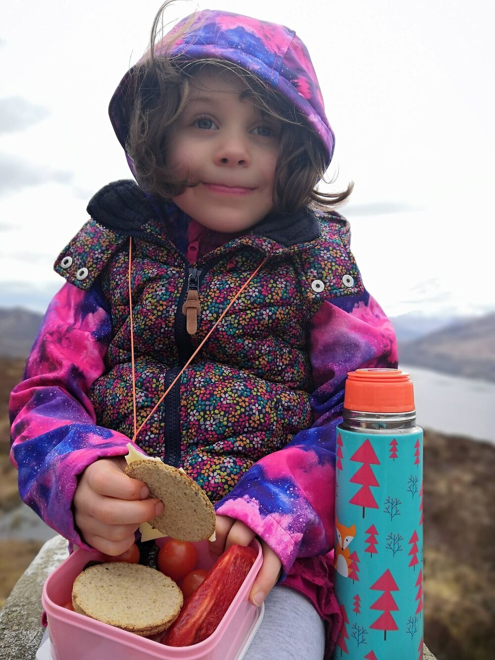 Small girl eating from a lunch box, with a loch behind her in the distance.