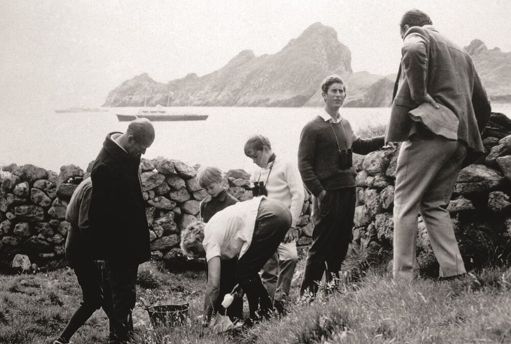 A black and white photograph of the Duke of Edinburgh, Prince Charles and other younger members of the royal family on St Kilda. Prince Charles stands by a stone wall, talking to a man with his back to the camera. The younger members of the party are looking at a hole in the ground with a lady carrying a trowel. The royal yacht is at anchor in the bay behind them.