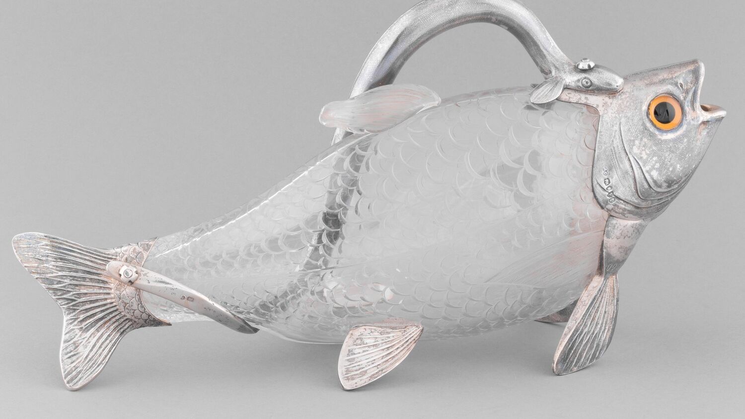 A hollow wine jug, shaped like a salmon, is made from glass and silver. It has a large yellow and black eye above the spout mouth. It is displayed against a plain grey background.