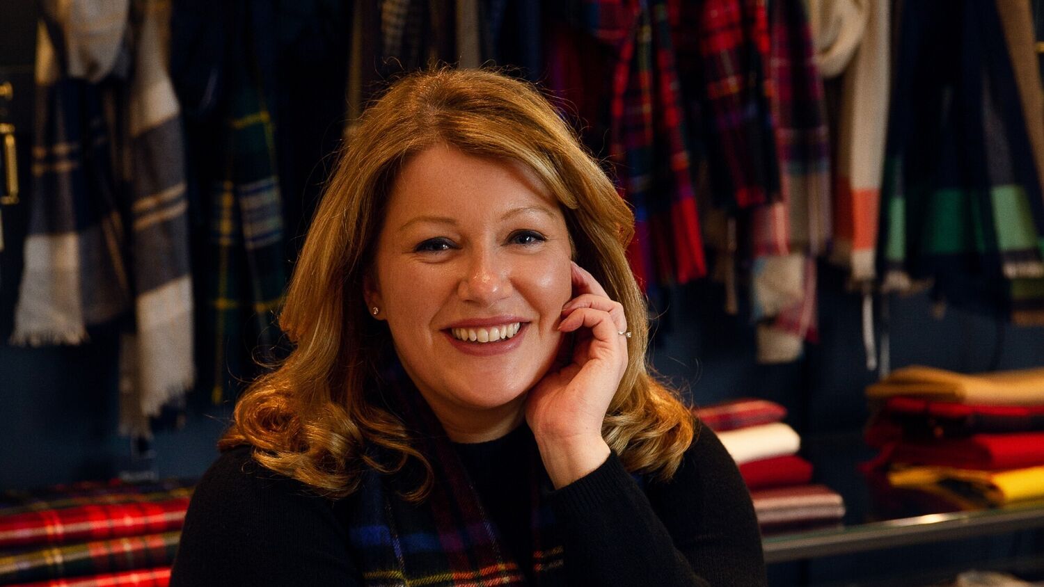 A smiling woman is sitting on a chair, wearing a tartan shawl.