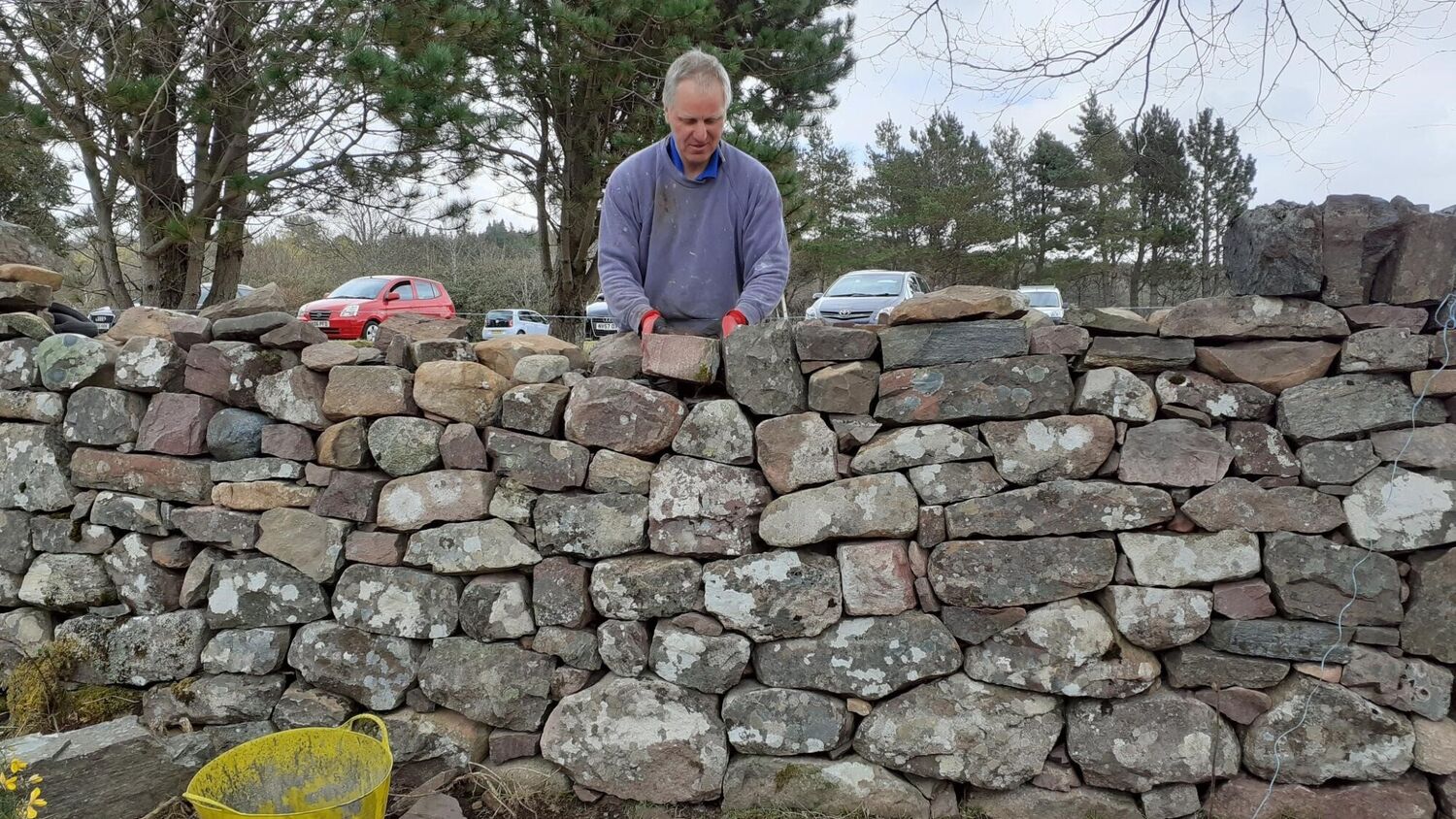A man is standing behind a drystone wall and is carrying out repairs to it.