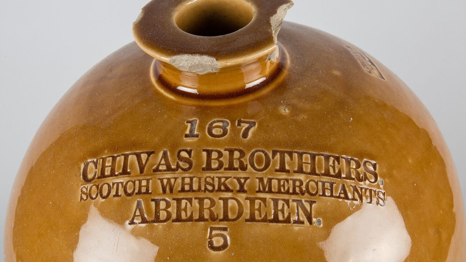 A close-up of the top half of an old whisky stoneware jar. It is very shiny and a mustard-yellow in colour. There are a couple of chips in the top spout. Printed on it is the following text: 167 Chivas Brothers. Scotch Whisky Merchants Aberdeen. 5.