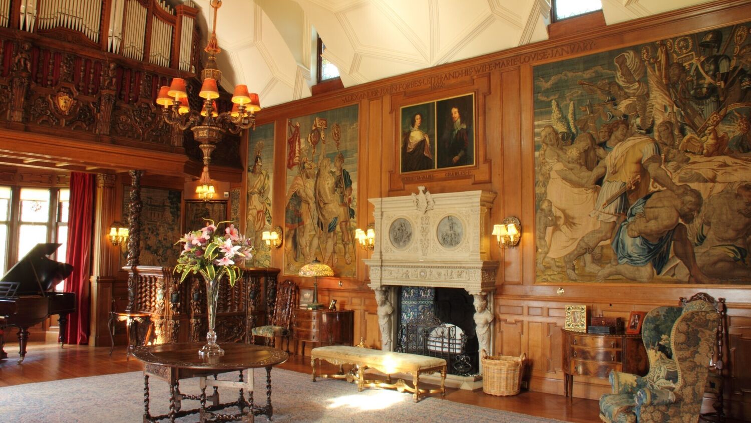 Grand room in a castle, with a large stone fireplace on a wood-panelled wall. There is a grand piano next to the window at one end of the room, and paintings and tapestries on the wall.