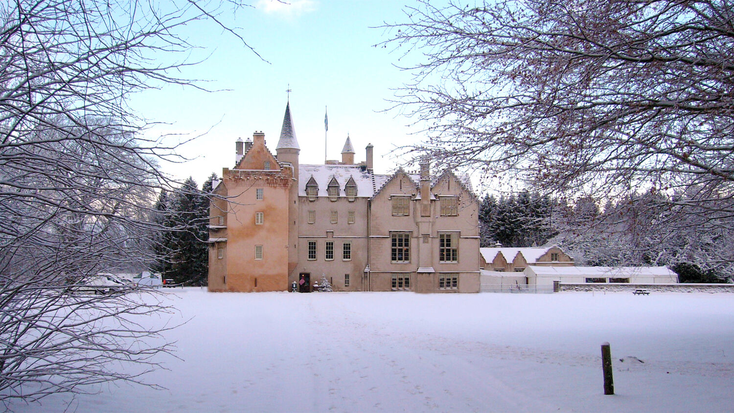 A view of Brodie Castle after heavy snowfall. The lawns and trees are blanketed with snow. The sky is a pale blue.