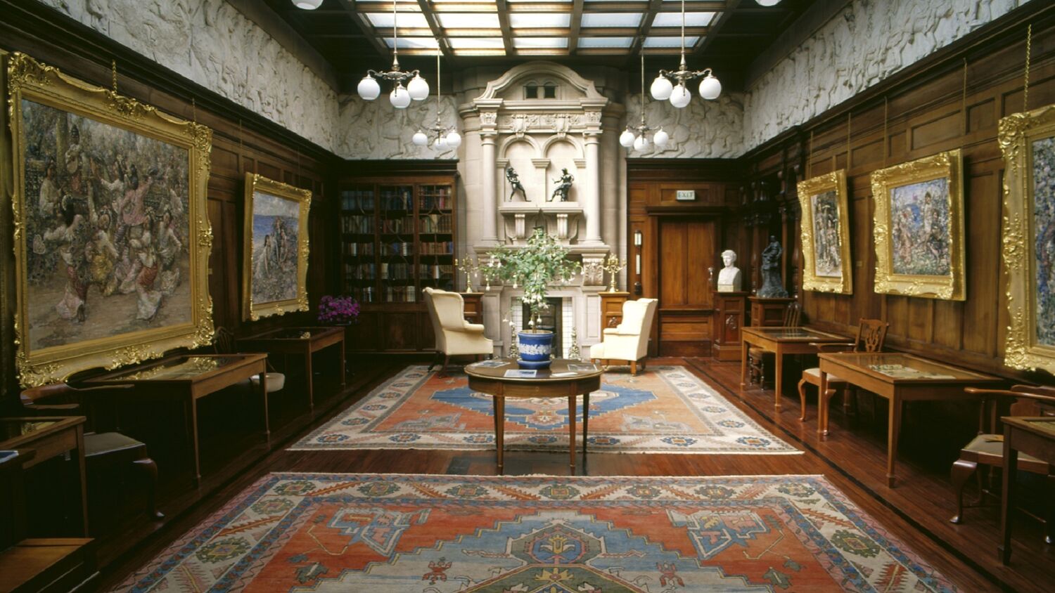 A view of a grand gallery room, with wood-panelled walls and skylight panels all along the ceiling. Gilt-framed paintings hang on the walls. The floor is covered by two large Turkish rugs. There is a very grand marble fireplace at the far end of the room, with two easy chairs before it. A number of wooden tables, some with vases of flowers upon them, stand around the room, with one in the centre.