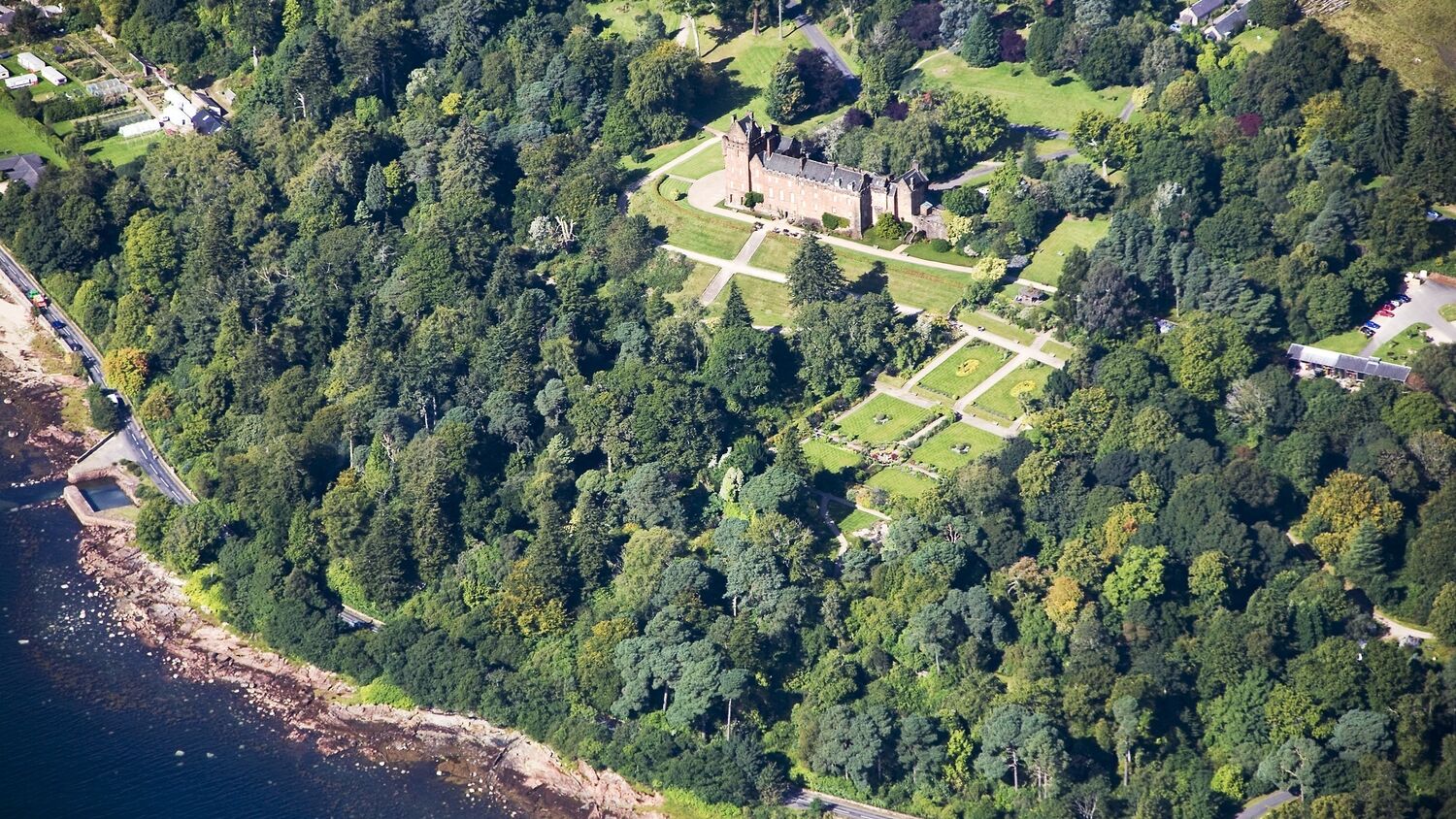 An aerial view of Brodick Castle, Garden and Country Park on Arran. The glistening blue bay can be seen in the bottom left corner. The castle can be seen with the garden in front, all surrounded by green woodland.