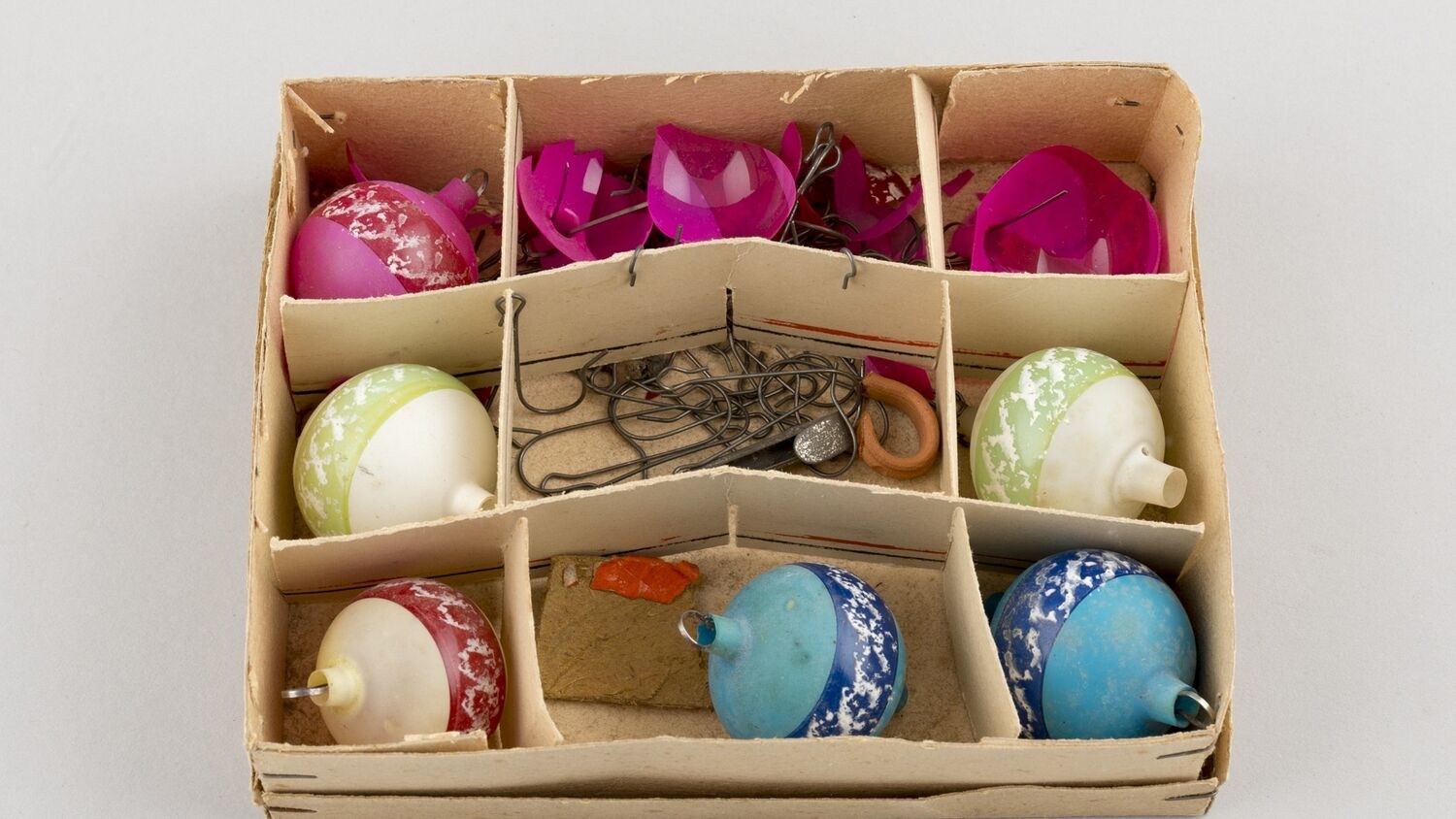 A brown cardboard box split into nine sections. In each of the section is a round Christmas bauble. Three are creamy white, three are hot pink and two are a bright duck-egg blue. Two of the pink baubles are shattered. The central section of the box is filled with metal pins.