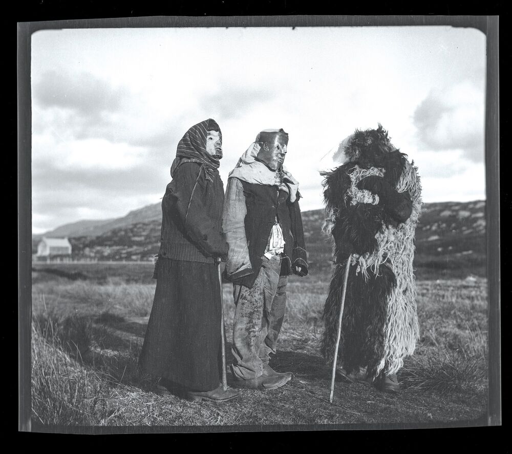 A black and white photograph of three people dressed in unusual Halloween costumes. The figure on the left wears a long dark skirt, a wool jacket and a mask made from a sheep's face. The figure in the centre has a tight leather mask tied over his face. The figure on the right is almost completely covered by a large, shaggy fleece, and carries a walking cane. All are standing outdoors in a field, with croft houses just visible in the background.