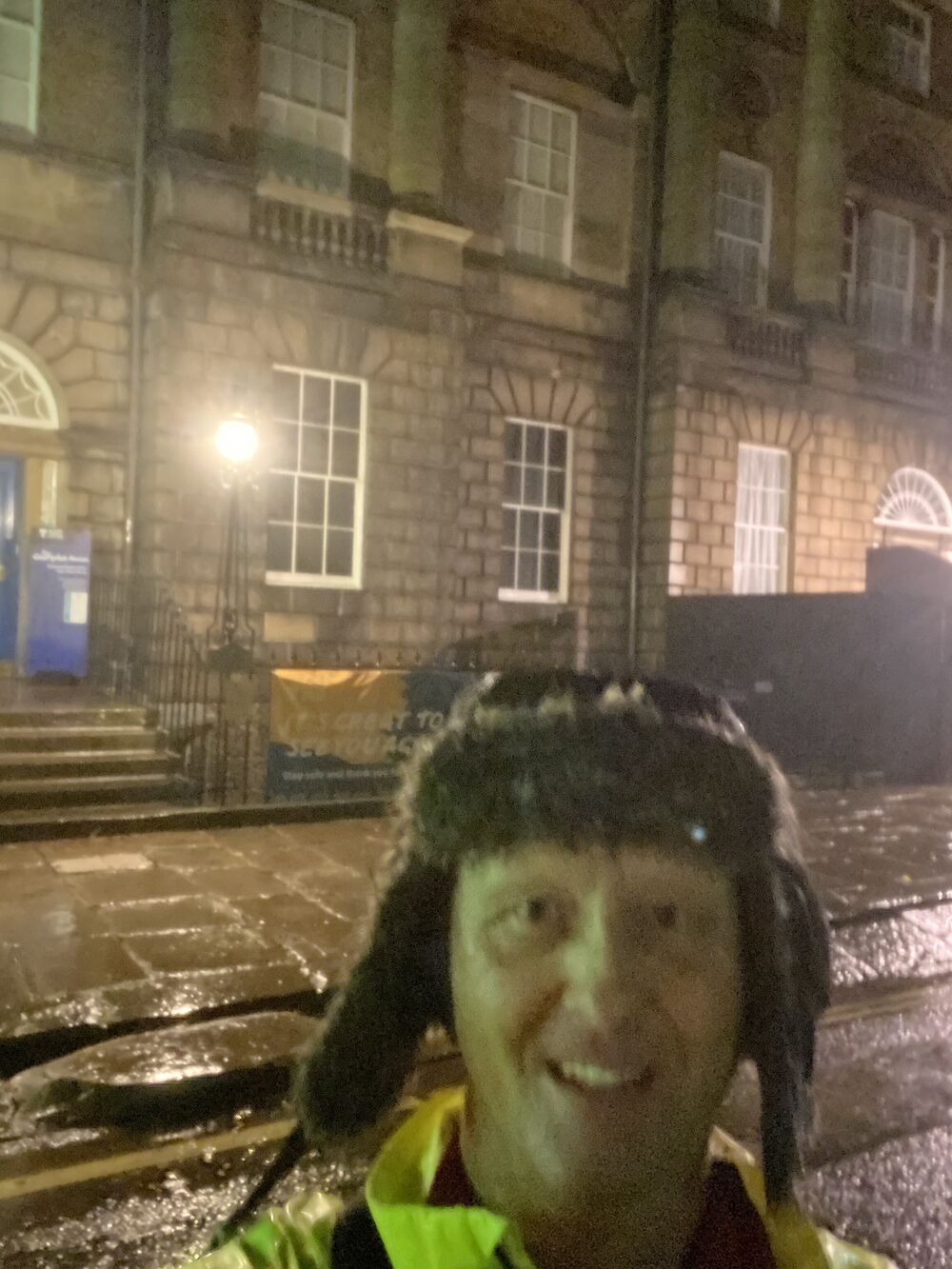 Man in hat and hi vis jacket standing in a rainy Edinburgh street in the dark