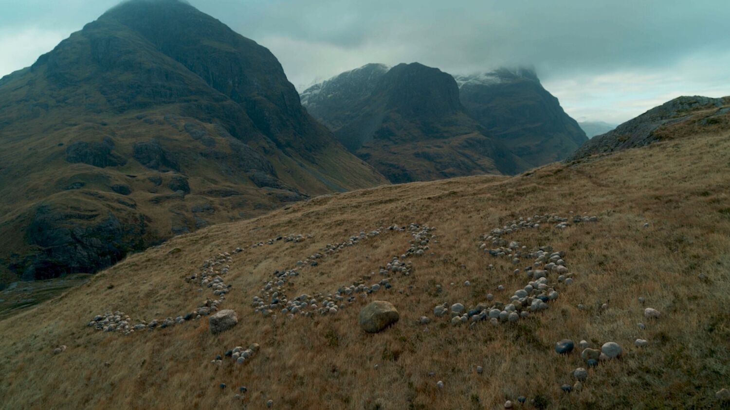 Moody image of Glencoe, with clouds over the mountain tops. In the foreground, on the hillside, are stones set out to spell SOS.
