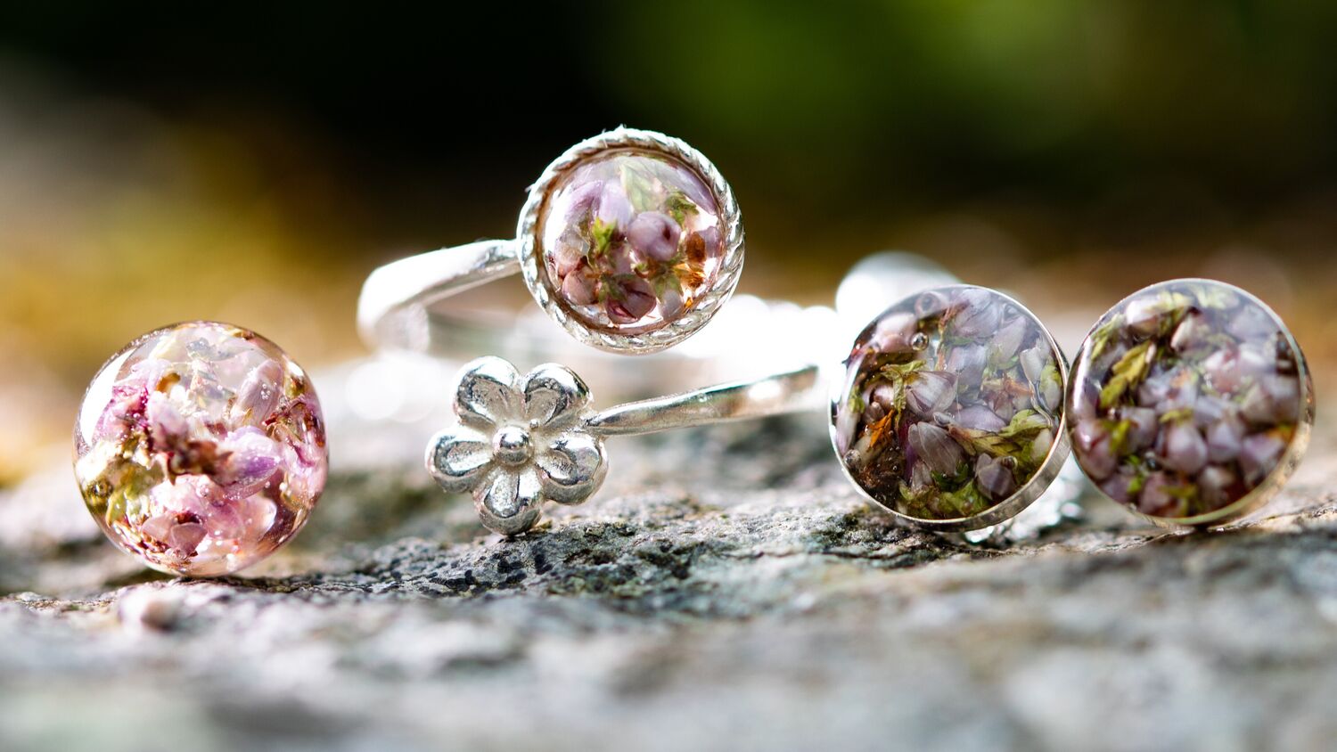 Close-up of a ring, pendant and earrings on a rock. The silver jewellery is made with heather embedded within resin.
