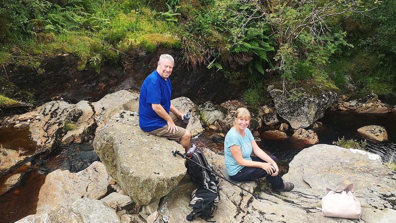A man and a woman sit on some large boulders at the edge of a burn, running down over rocks. Both turn towards the camera and smile. A large rucksack and a walking pole lies on the rock between them, with a smaller rucksack at the woman’s feet. The man is holding a water bottle.