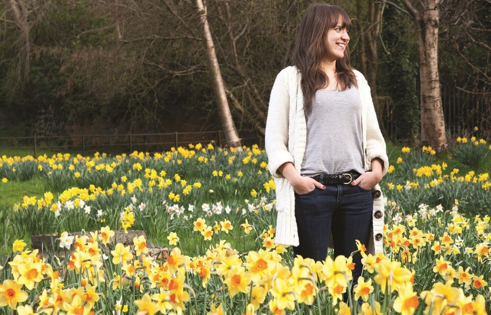 A lady stands in a garden, surrounded by beds of different varieties of daffodils.