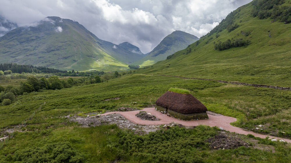 A reconstructed cottage with a heather-thatched roof stands in a clearing in a glen. Tall mountains rise all around it, with heavy clouds passing over the tops.