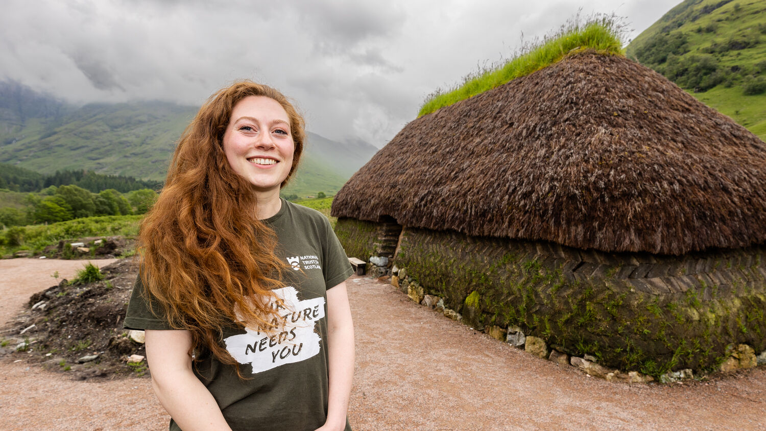 Woman in brown t-shirt with long red hair stands next to traditional thatch longhouse