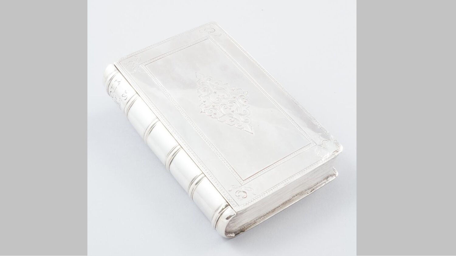 A little silver oblong box, shaped like an old book, is displayed against a pale grey background. It has some etched patterns on the top and side.