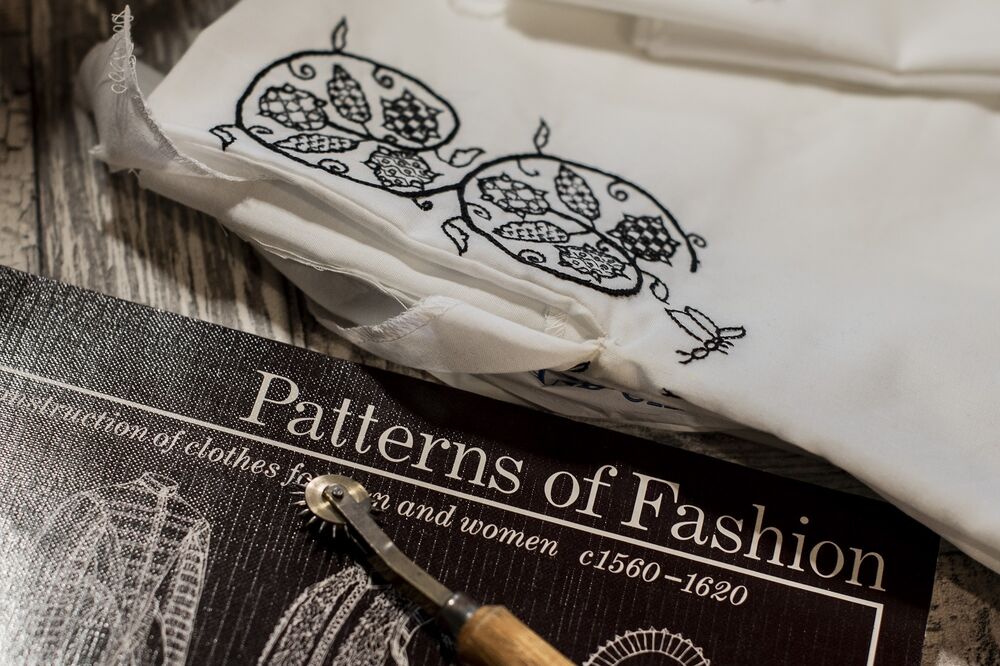 A piece of white fabric, with intricate black embroidered patterns, lies on a desk beside a book called Patterns of Fashion. A cloth-cutting wheel lies on top of the book.