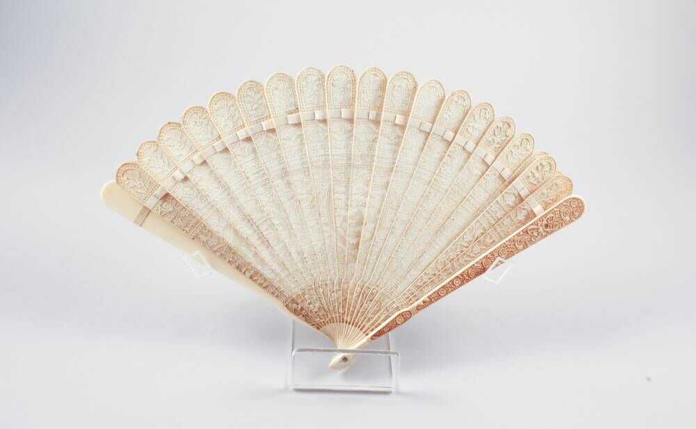 An ivory fan with tiny detailing on each individual leaf, propped up in a clear holder.