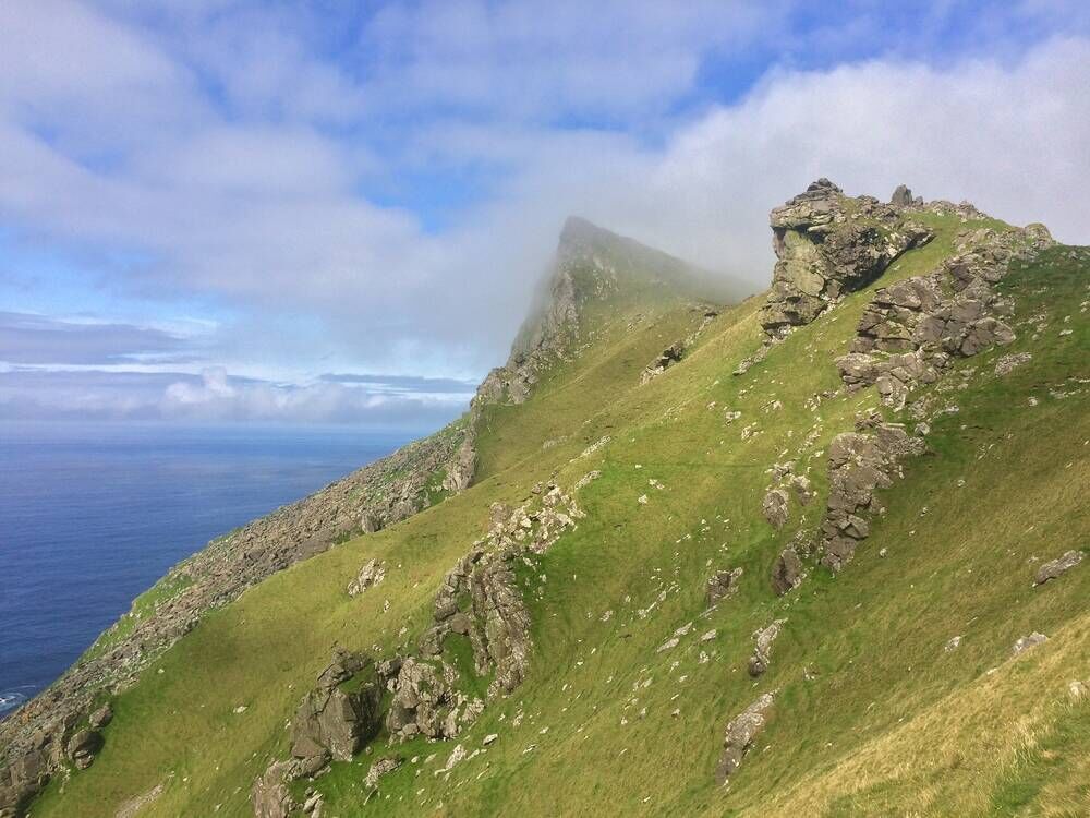 A view of an incredible steep ridge on an island, plummeting straight down towards the sea. The sides are grassy, with rocky crags nearer the top. Light clouds hug the top of the crags.