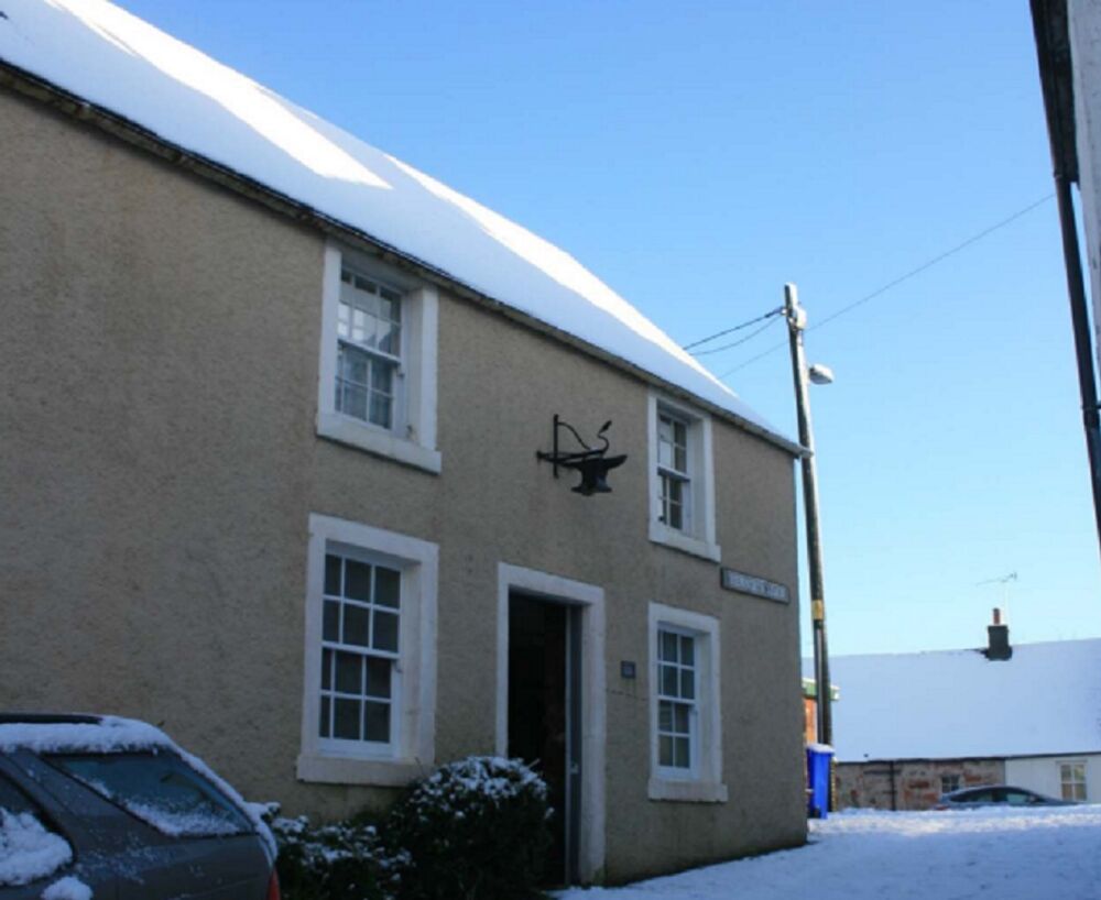 An end of terrace house on a narrow street. Snow covers the road and roofs. A car is parked outside the house. There is a metal sculpture protruding from the wall above the main door.
