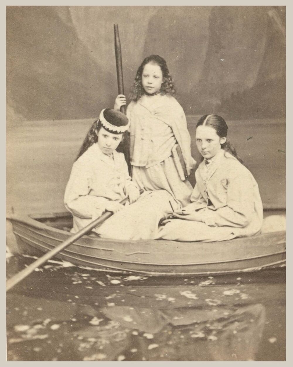A sepia photograph of three children sat in a rowing boat. Each girl has dark hair and is wearing a white dress. One sits holding an oar, one sits in front of her, and the other stands behind them.