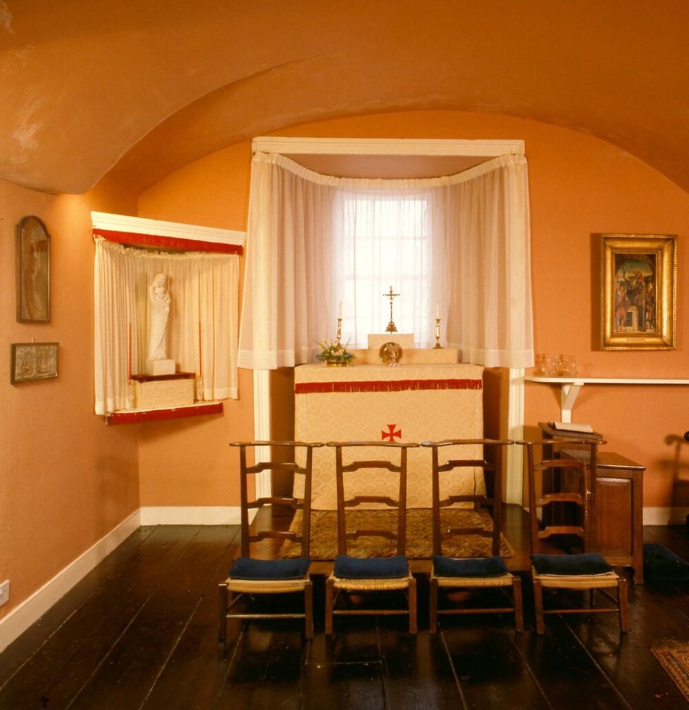 Four wooden chairs are lined up in a row, in a chapel room with an arched ceiling painted in a sunny pale orange. Religious pictures, crosses, and a small white statue of the Virgin Mary line the walls, windowsill and a small curtained alcove in the corner.