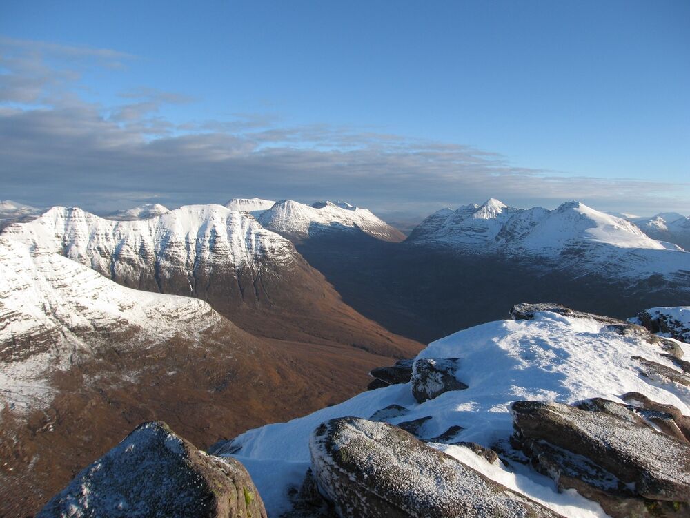 A view from a mountain ridge, looking across at many other tall snow-covered peaks. A river can just be seen snaking through the barren brown glen far below.