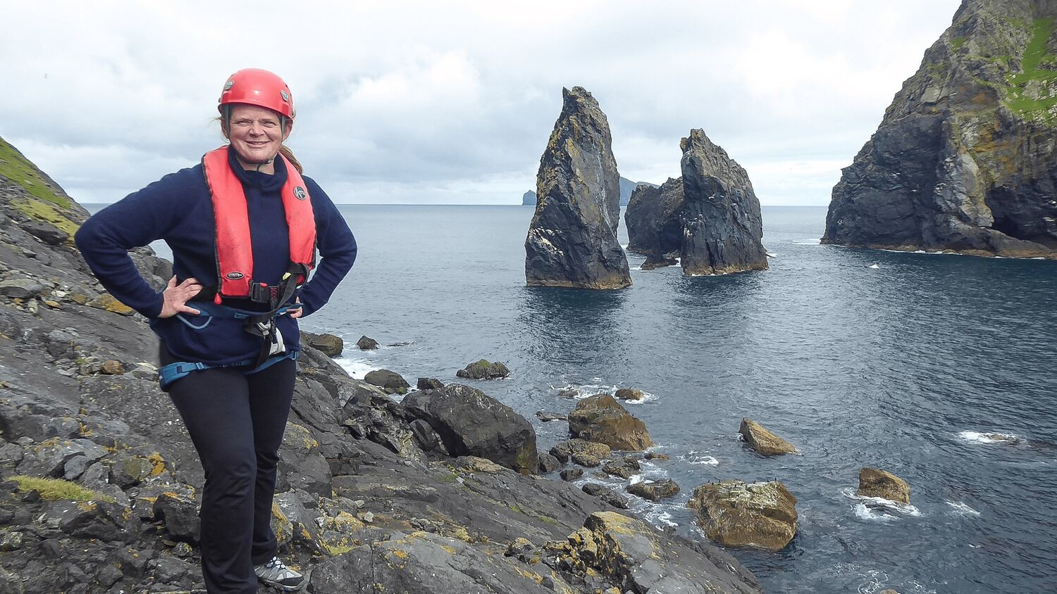 A woman wearing a red helmet and red floatation jacket stands on a rocky ledge beside a sea inlet. Large rock stacks stand just off shore behind her.