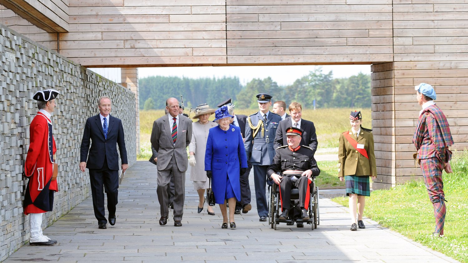 Her Majesty The Queen, the Duke of Edinburgh and a large delegation of official people walk along the wide path outside the new visitor centre at Culloden. The Queen is in the centre, wearing a blue hat and dress.