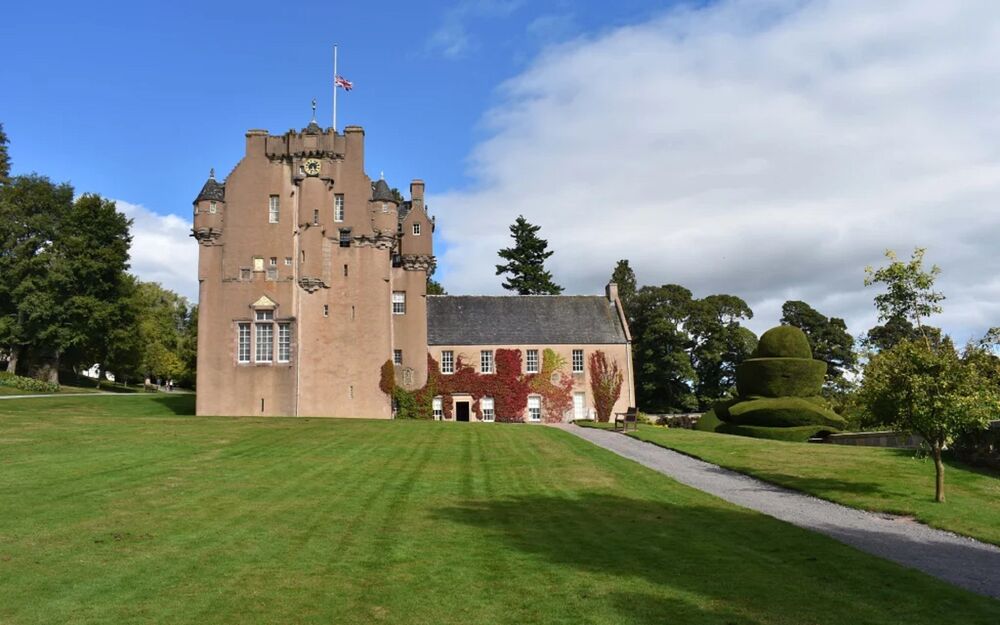 A Union Jack flag flies at half-mast on Crathes Castle, fluttering against a blue sky with white clouds.