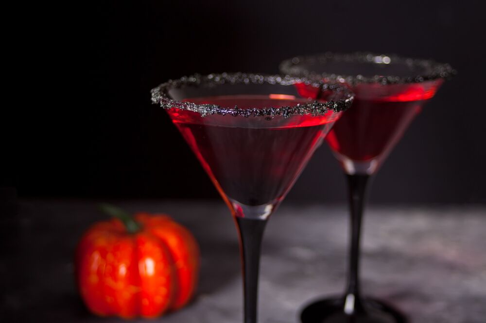 Two martini glasses are filled with a deep red cocktail. A bright orange pumpkin lies on the table behind them.