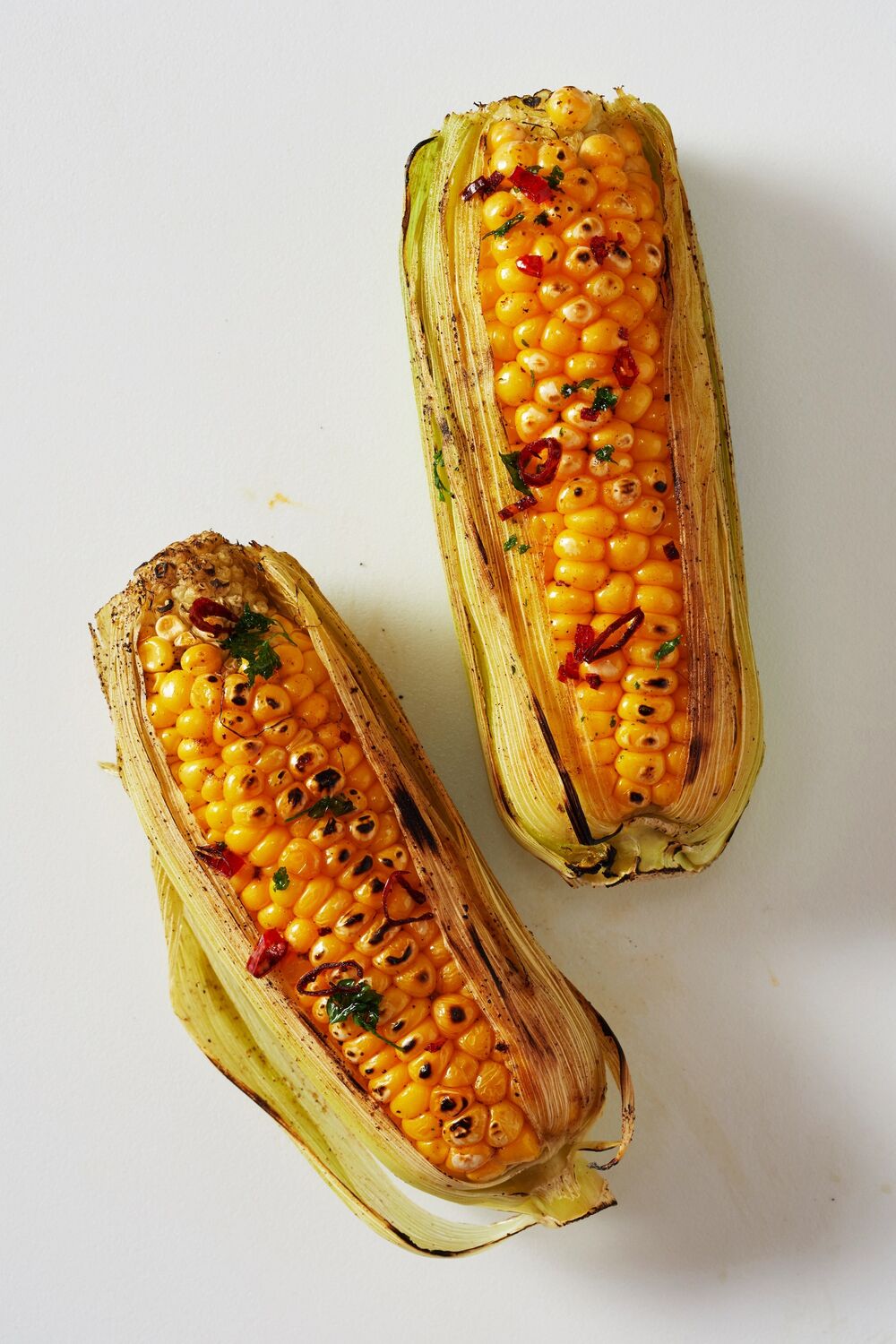 Two corn on the cobs lie side by side on a plain surface. They still have their leaves enveloping them. They have been grilled with butter and spices.
