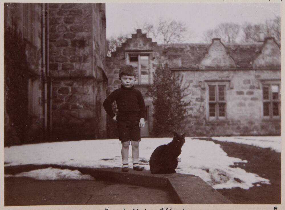A black and white photo of a small boy and a black cat standing on a step outside a castle. Snow lies on the ground in the background.