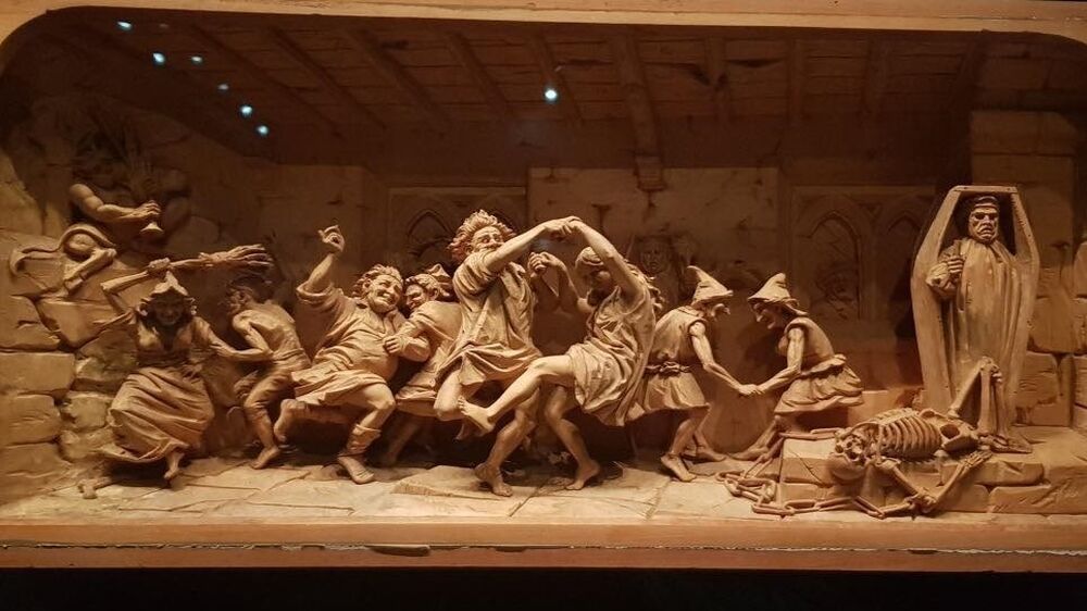 A miniature carved wood 3D scene depicts figures dancing and cavorting in an old church. A skeleton is chained to the floor.